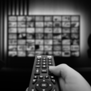 A first-person black and white view of a hand holding a television remote control pointed at a large, blurred screen displaying a grid of streaming media content.